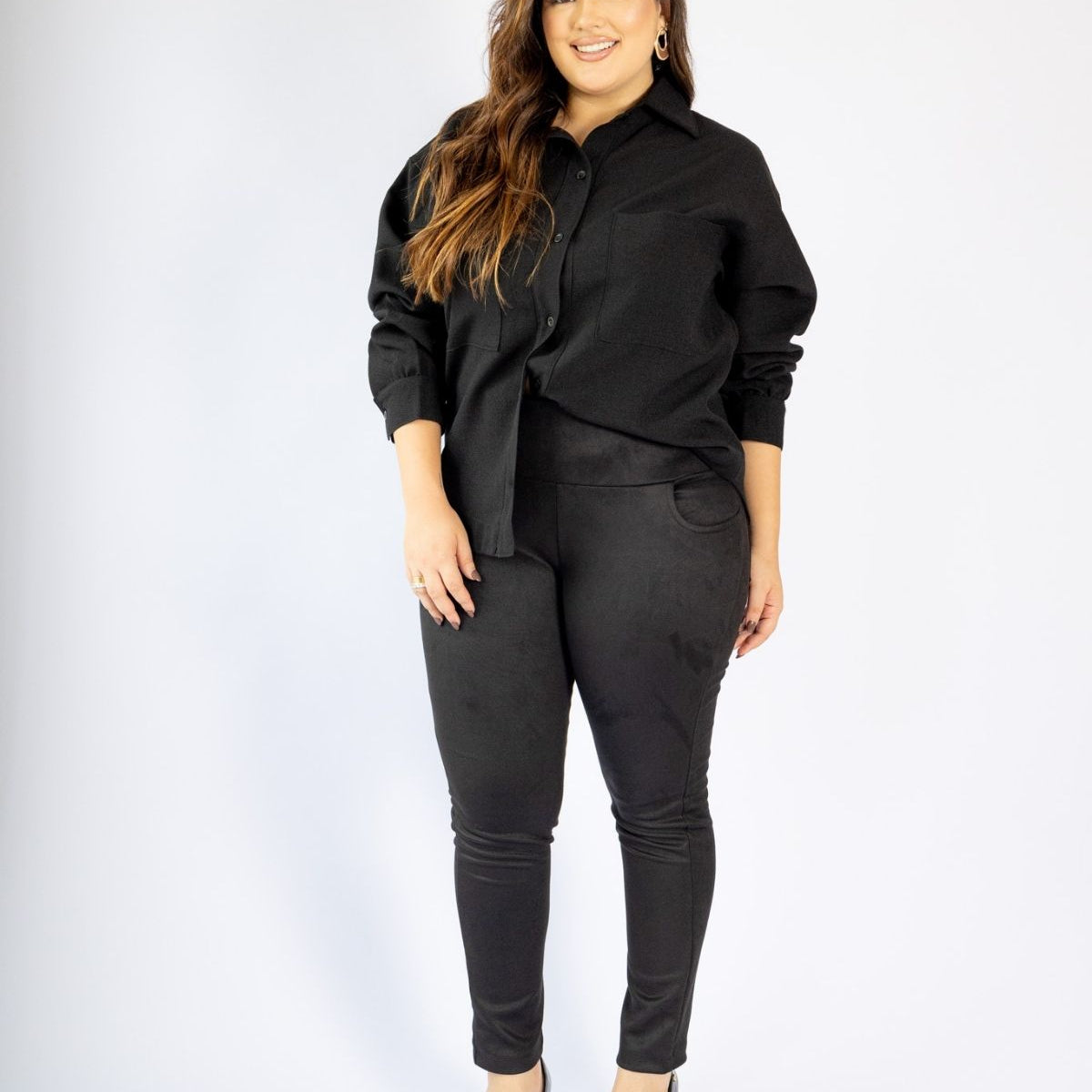 Mulher usando conjunto modelador preto com calça skinny e camisa, em fundo branco, moda plus size.
