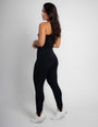 Mulher usando calça modeladora preta de cintura alta e top preto, vista traseira, fundo neutro.
