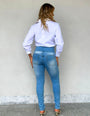 Mulher de costas vestindo calça jeans modeladora cintura alta e camisa branca, fundo neutro.